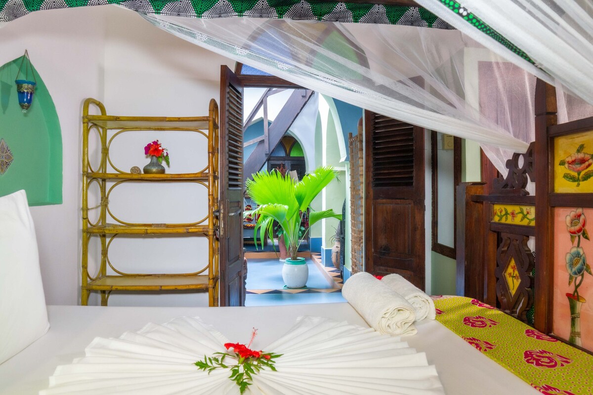 A room with a bed, a bamboo shelf with a vase, a potted plant, and a doorway leading to another room.