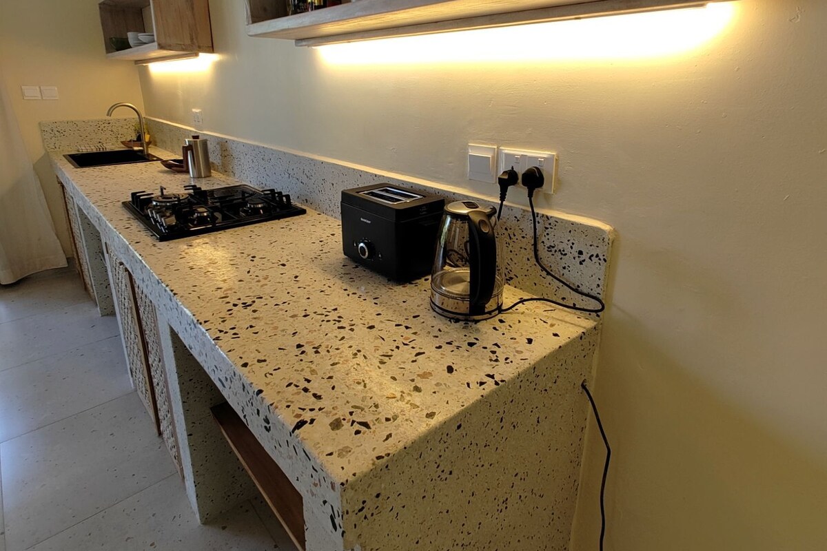 A kitchen with a speckled countertop, a built-in gas stove, a toaster, and a coffee maker. There is a wooden shelf above the countertop with a few items on it.