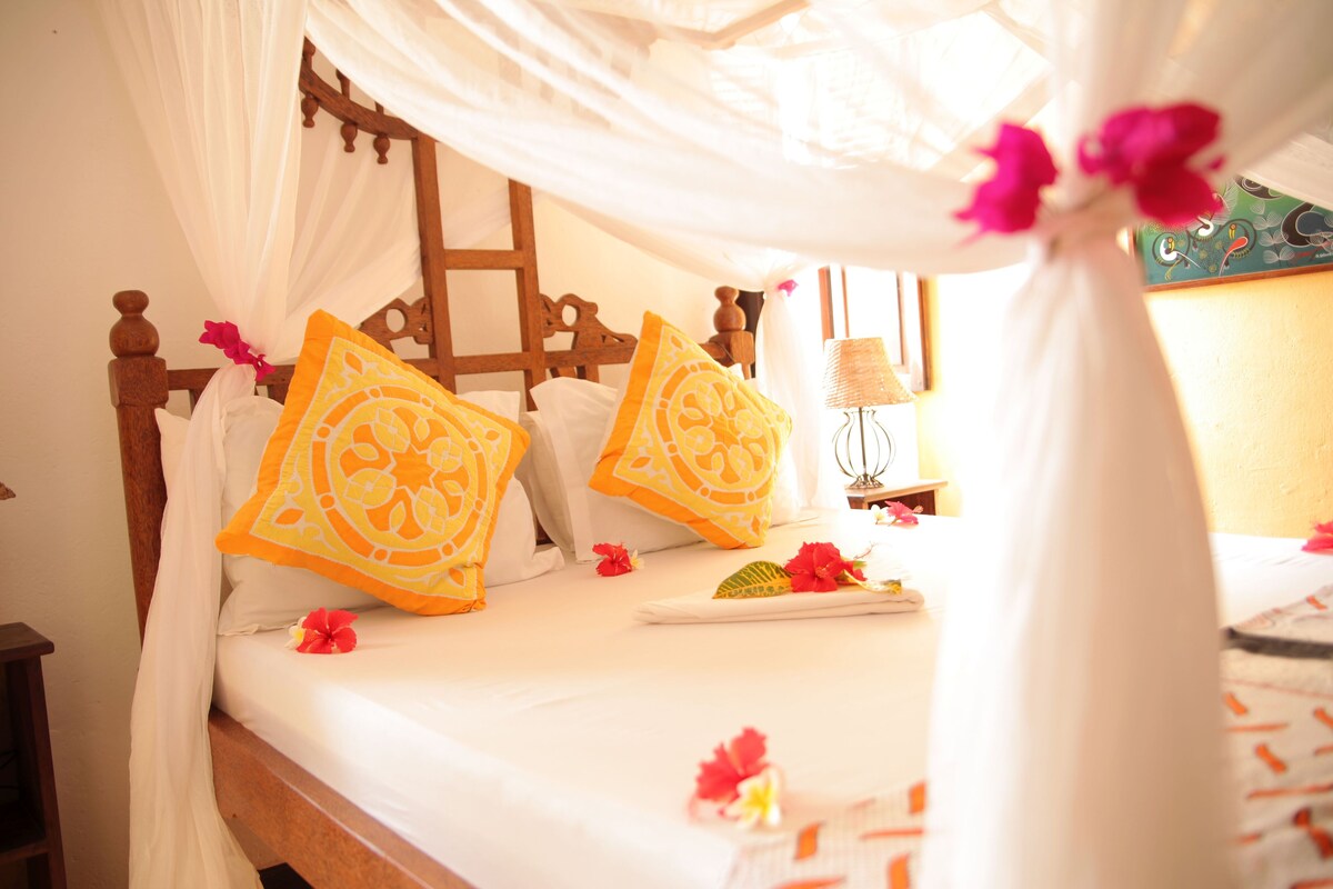 A bed with a canopy, two orange patterned pillows, and a bedside table with a lamp.