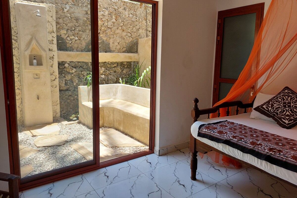 A bedroom with a bed, a wooden bench, a stone wall, and a glass door leading to an outdoor area with a bench and plants.