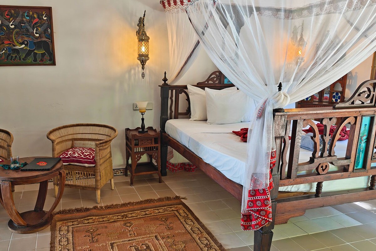 A room with a bed, two wicker chairs, a small table, a decorative rug, a wall hanging, and a nightstand with a lamp.