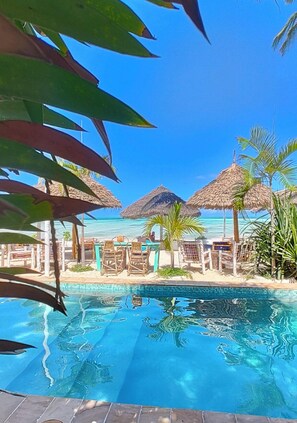 A clear blue pool with thatched umbrellas and lounge chairs, surrounded by palm trees and a view of the ocean. - Villa Bahati Zanzibar (Jambiani)