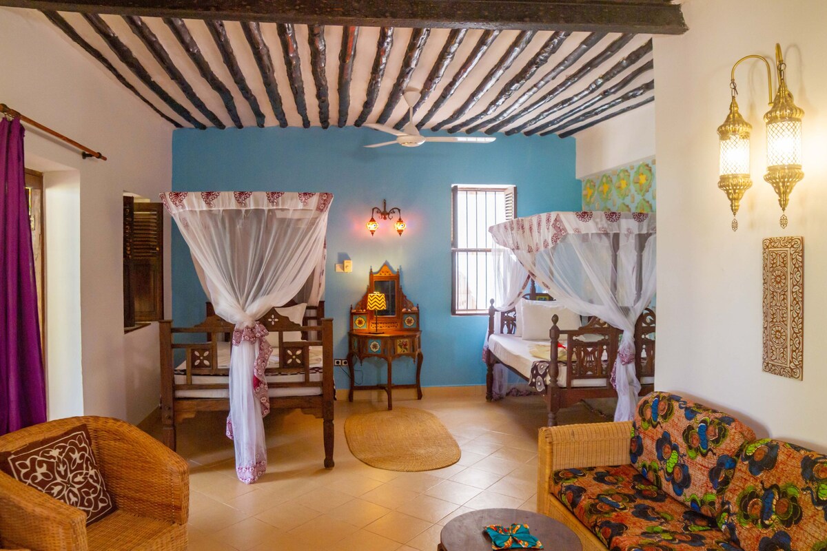 A room with a blue wall, wooden ceiling beams, a ceiling fan, two chairs with canopy beds, a sofa, and a small table.