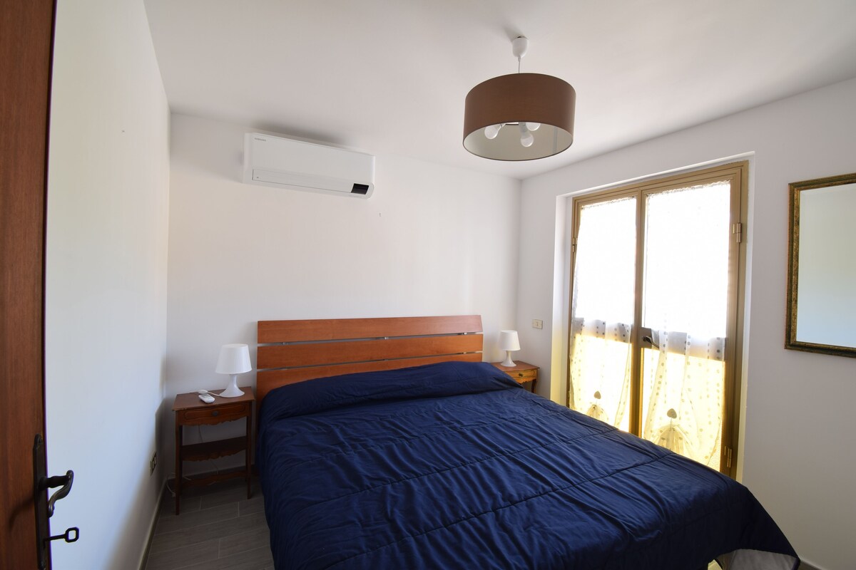 Ein Schlafzimmer mit einem hölzernen Kopfteil, einem Bett mit blauer Bettdecke, einem Nachttisch mit Lampe und einem Fenster mit Vorhängen.
