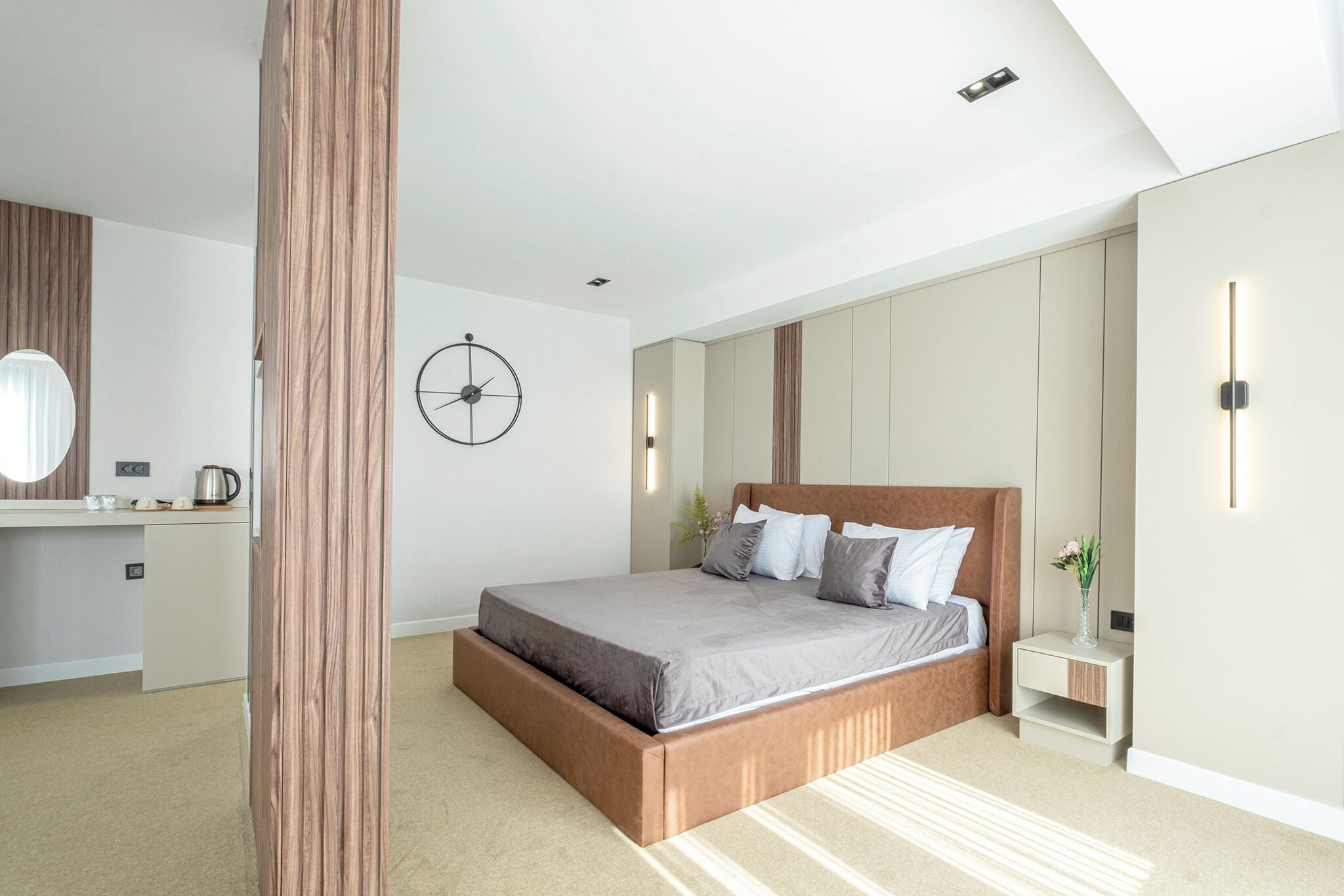 A modern bedroom with a large bed, a bedside table, a nightstand, a wall clock, and a built-in wardrobe.