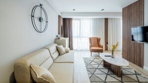 A modern living room with a beige sofa, a coffee table, a TV mounted on the wall, and a large window with curtains. - EUPHRATES HOTEL (Bursa)