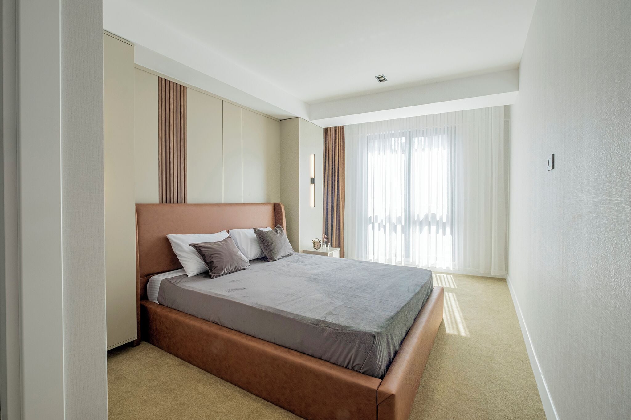 A modern hotel room with a large bed, neutral color walls, and a large window with sheer curtains.