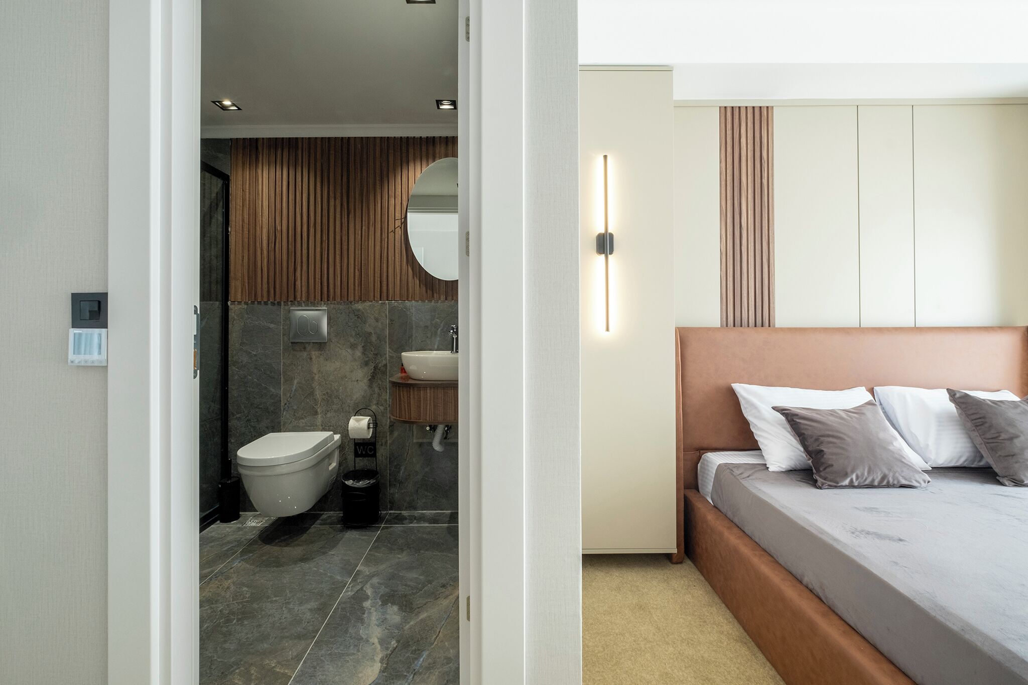 A modern bathroom with a toilet, sink, and mirror, and a bedroom with a bed and bedside table.