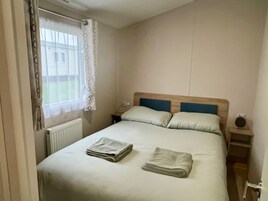 Un dormitorio con cama, dos almohadas, una mesita de noche, lámpara y ventana con cortinas.