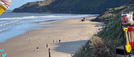 A beach with a cliff in the background, a few people walking, and a surfboard rack on the right.