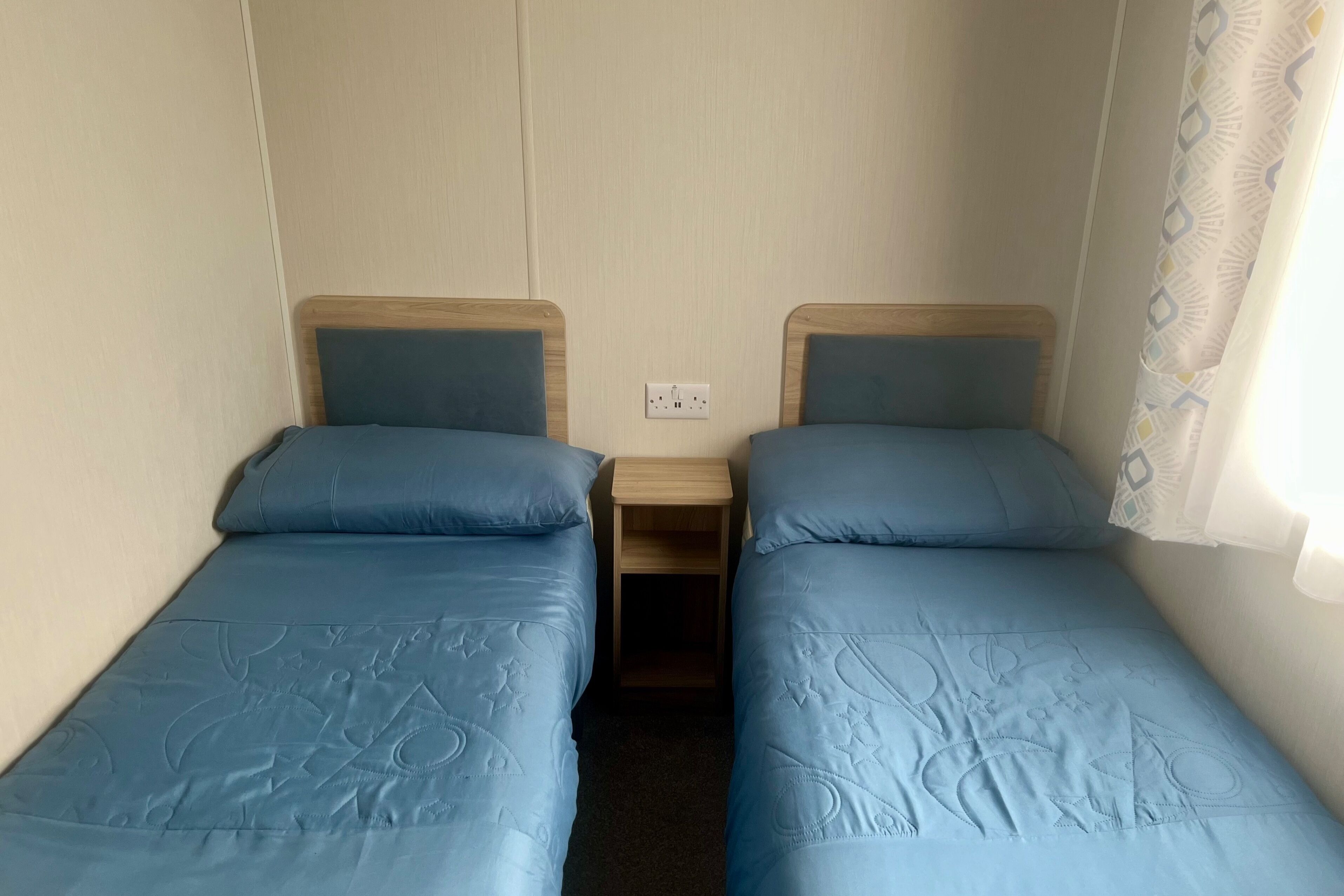 Une chambre avec deux lits simples, chacun doté d’une tête de lit en bois et d’un matelas bleu assorti. Une petite table de chevet en bois se trouve entre les lits, et une prise de courant est fixée au mur.