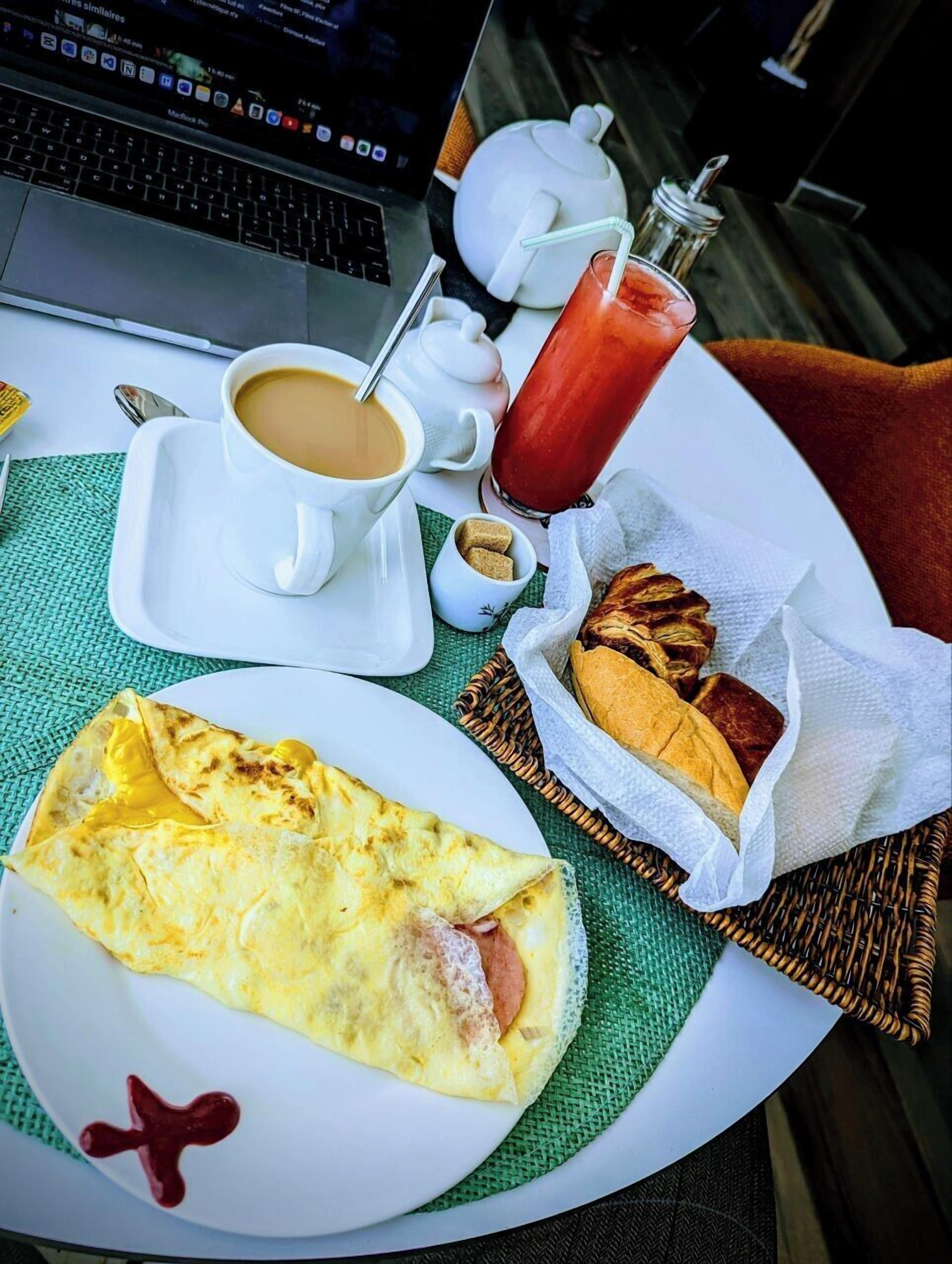 Un desayuno con laptop, café, una bloody mary, pan y una tortilla en una mesa.