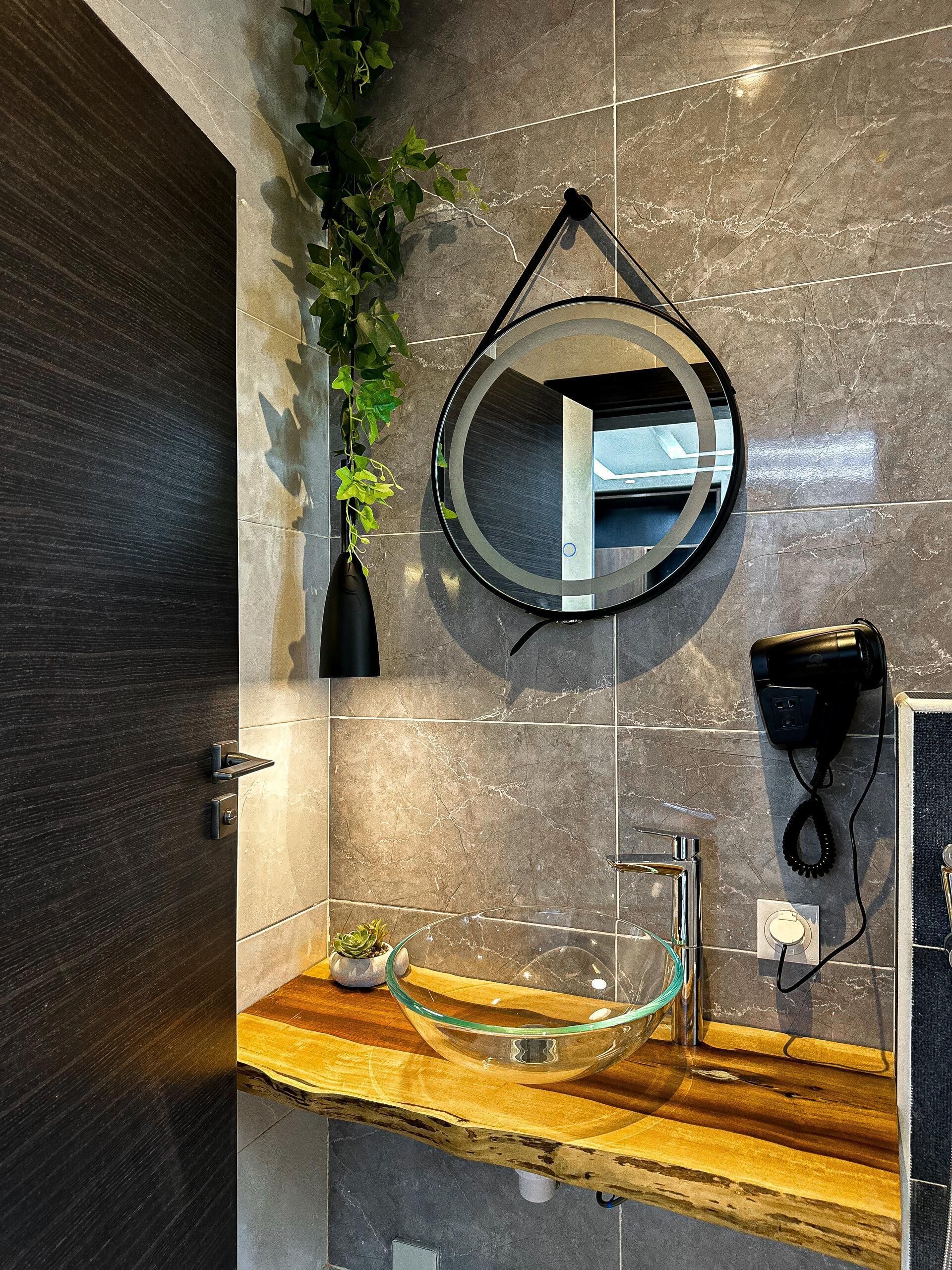 Een badkamer met een glazen wasbak, een ronde spiegel, een haardroger en een potplant.