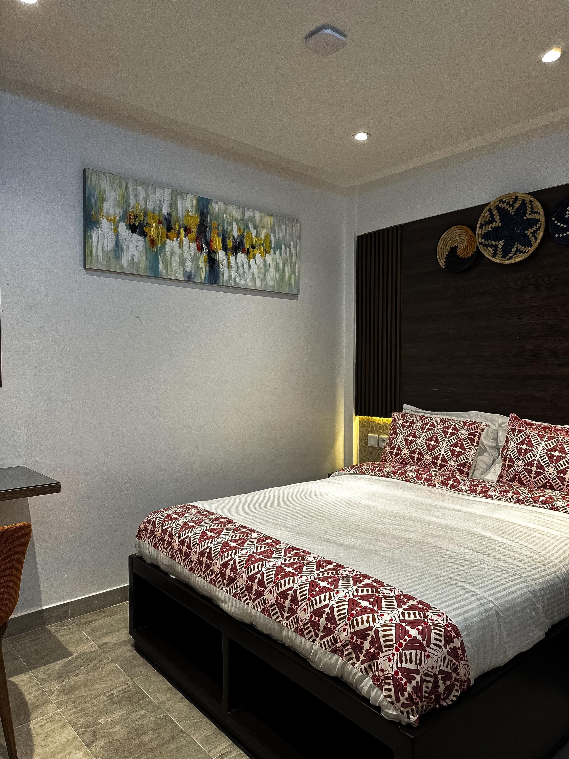 A modern bedroom with a large bed, a desk, and a wall-mounted abstract painting.