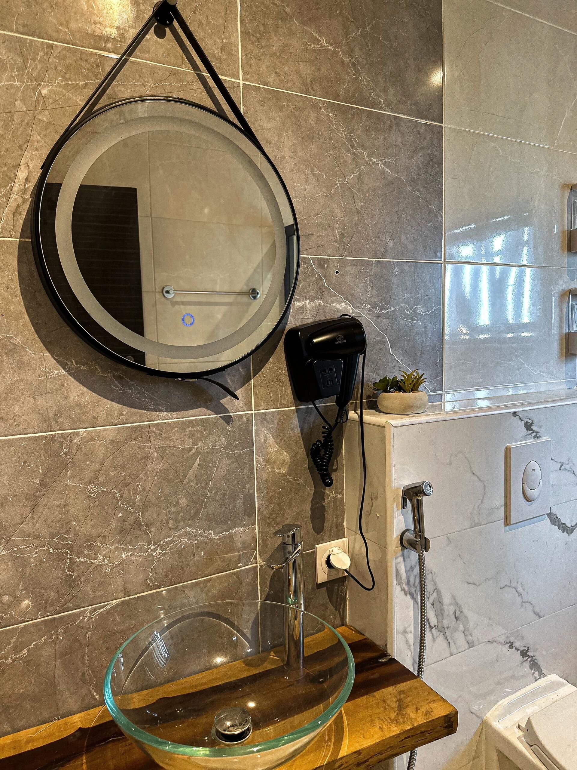 A bathroom with a round mirror, a glass sink, a handheld hair dryer, and a shower.