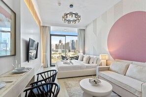 Een moderne hotelkamer met een groot raam dat uitzicht biedt op de stad, een witte bank, een zithoek met een tafel en stoelen, en een sierlijke kroonluchter.