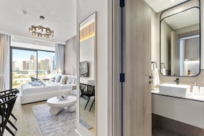 Een moderne hotelkamer met een groot raam, een bank, een zithoek en een badkamer met een grote spiegel en wastafel.