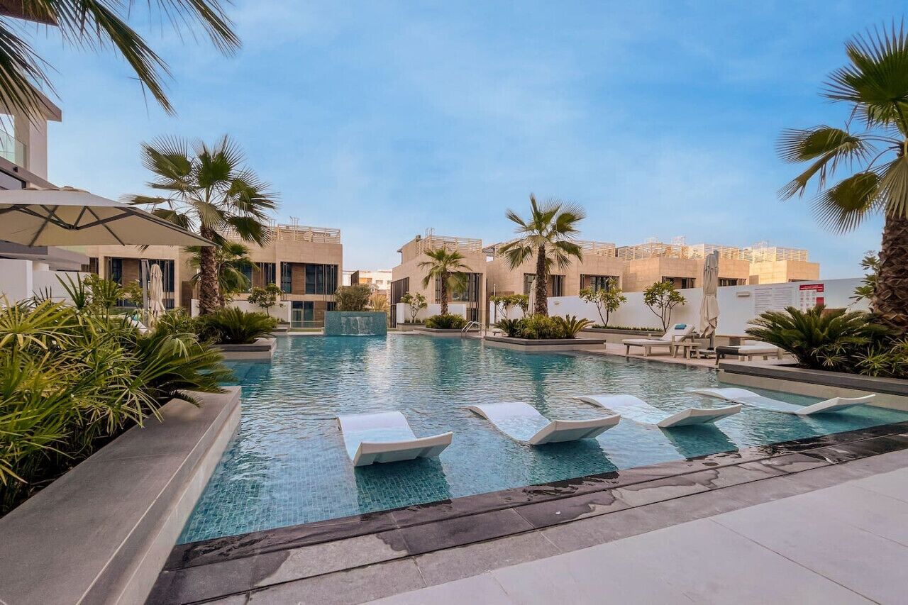 A swimming pool with lounge chairs, palm trees, and modern buildings in the background.