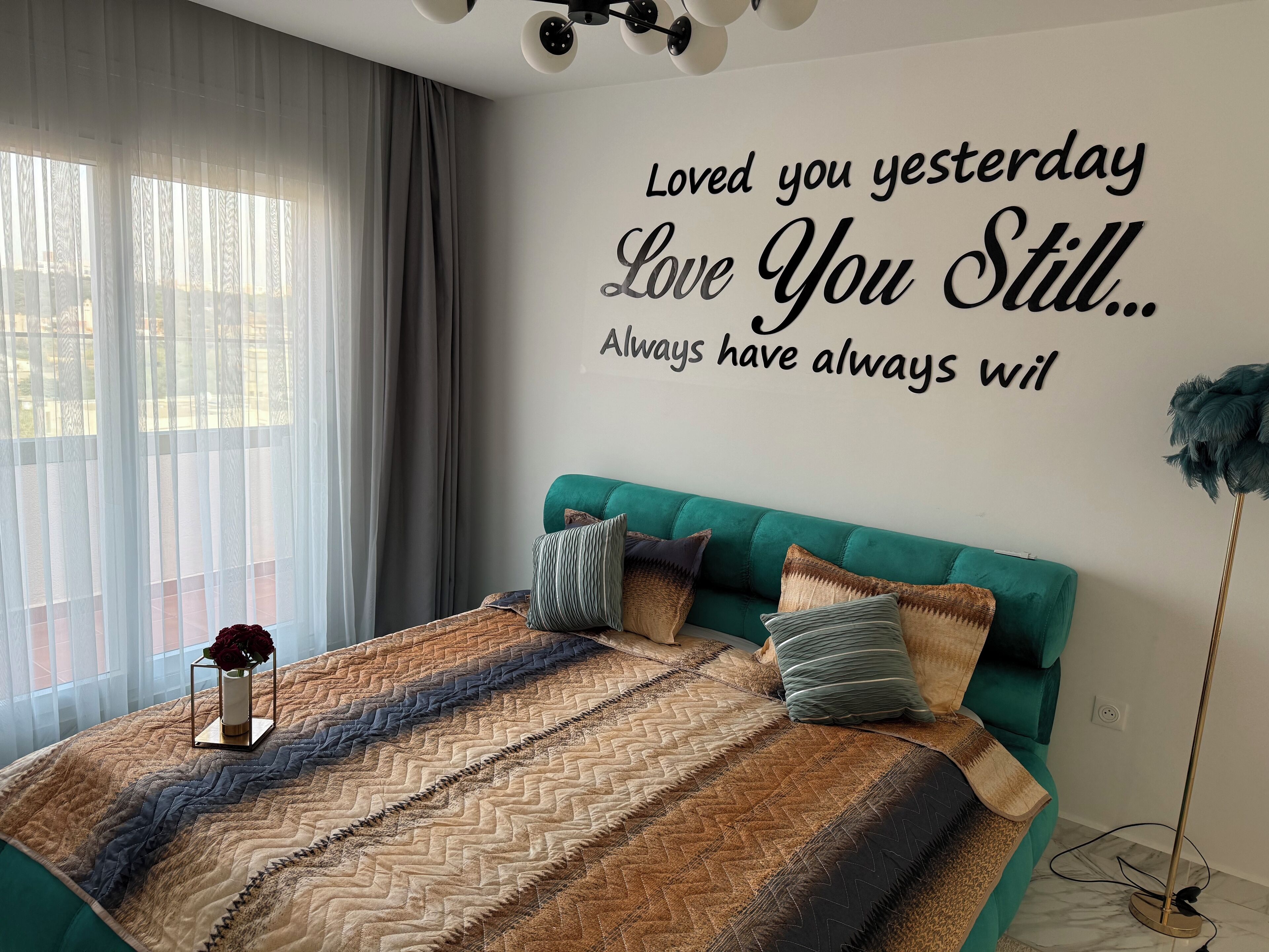 Un dormitorio con cama, cabecera color turquesa, cita decorativa en la pared y un jarrón con una flor.