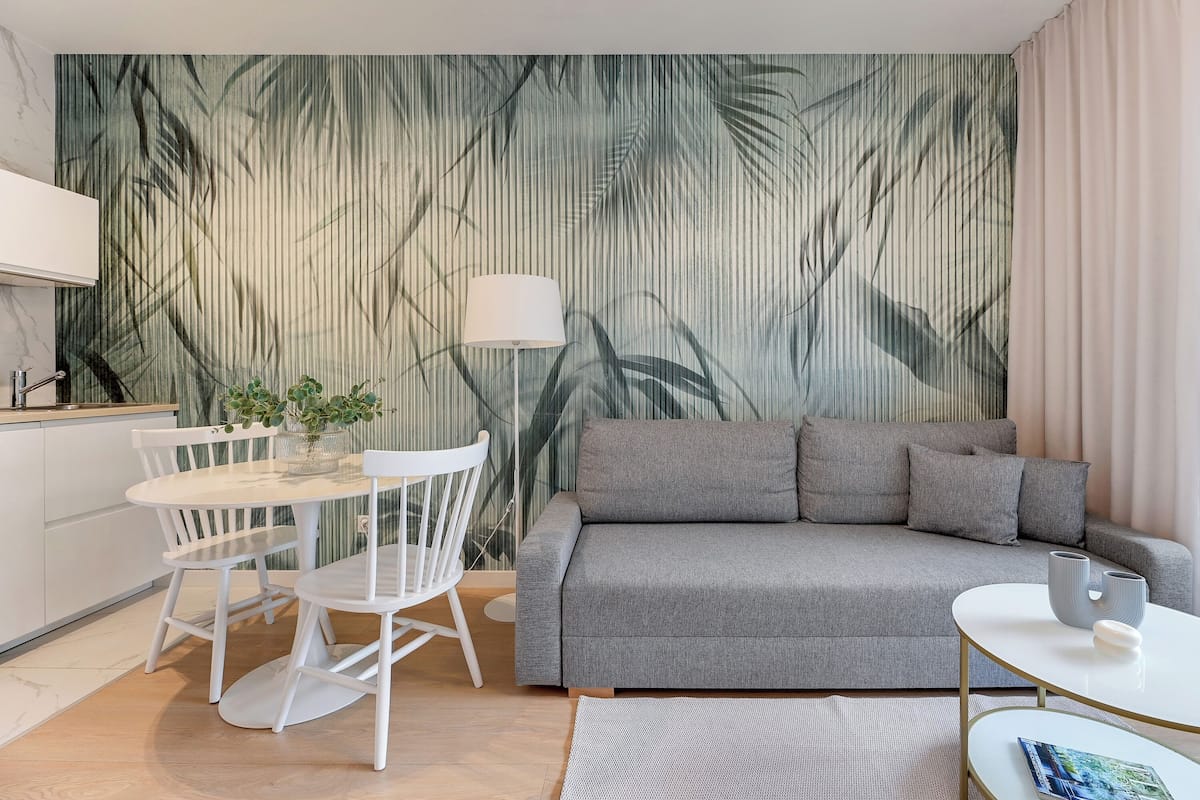 Un salon moderne avec un canapé gris, des chaises blanches, une petite table et un mur orné d’un motif de feuilles tropicales.