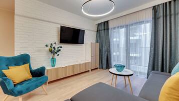 Ein modernes Wohnzimmer mit weißer Ziegelwand, einem Flachbildfernseher, einem blauen Sessel, einem grauen Sofa, einem kleinen runden Tisch und einem großen Fenster mit Vorhängen.