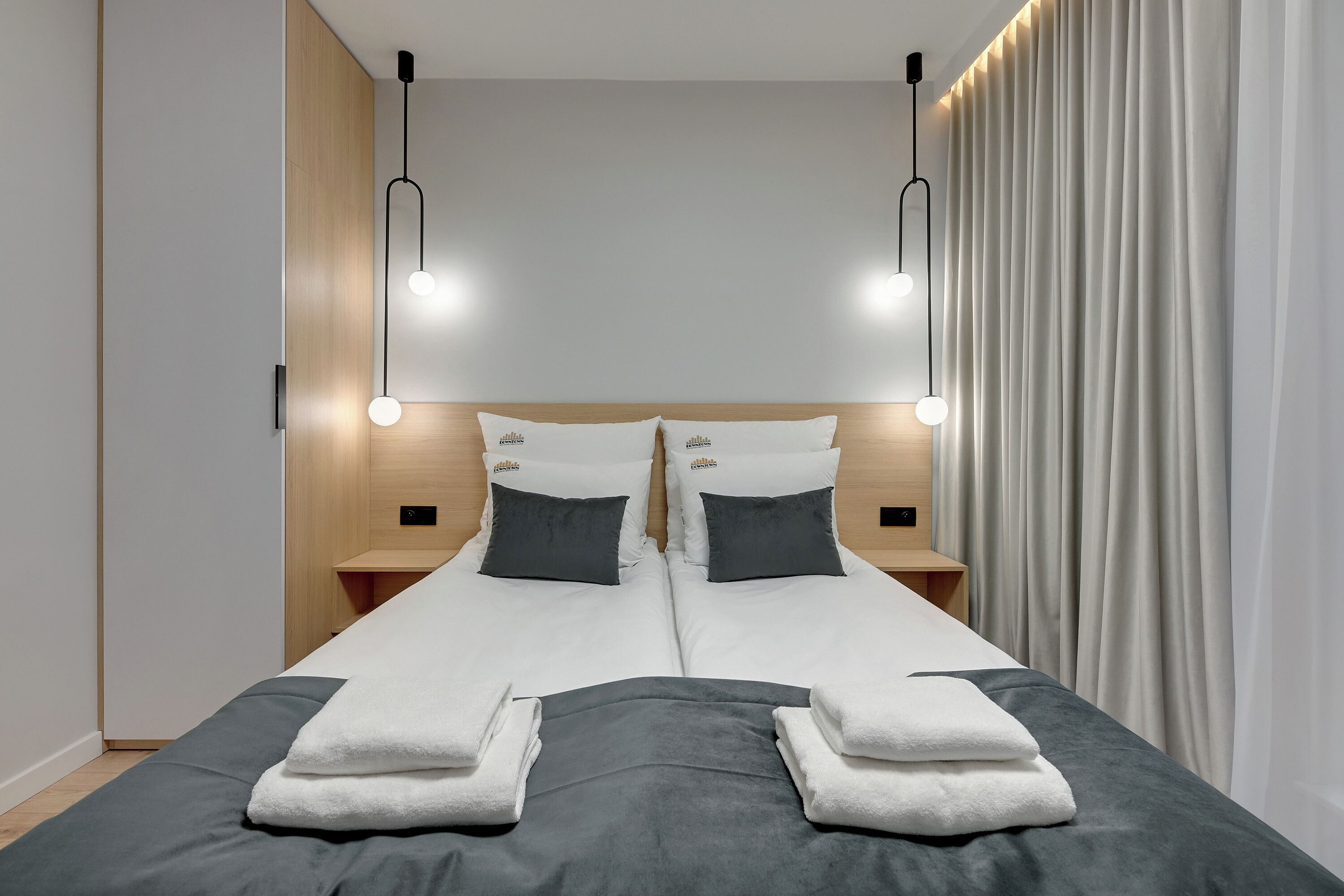 Un lit double avec une literie blanche et grise, deux tables de chevet avec des lampes et un rideau sur le côté droit.
