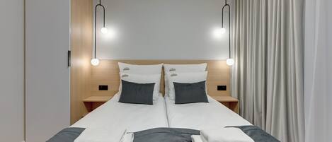 Un lit double avec une literie blanche et grise, deux tables de chevet avec des lampes et un rideau sur le côté droit.