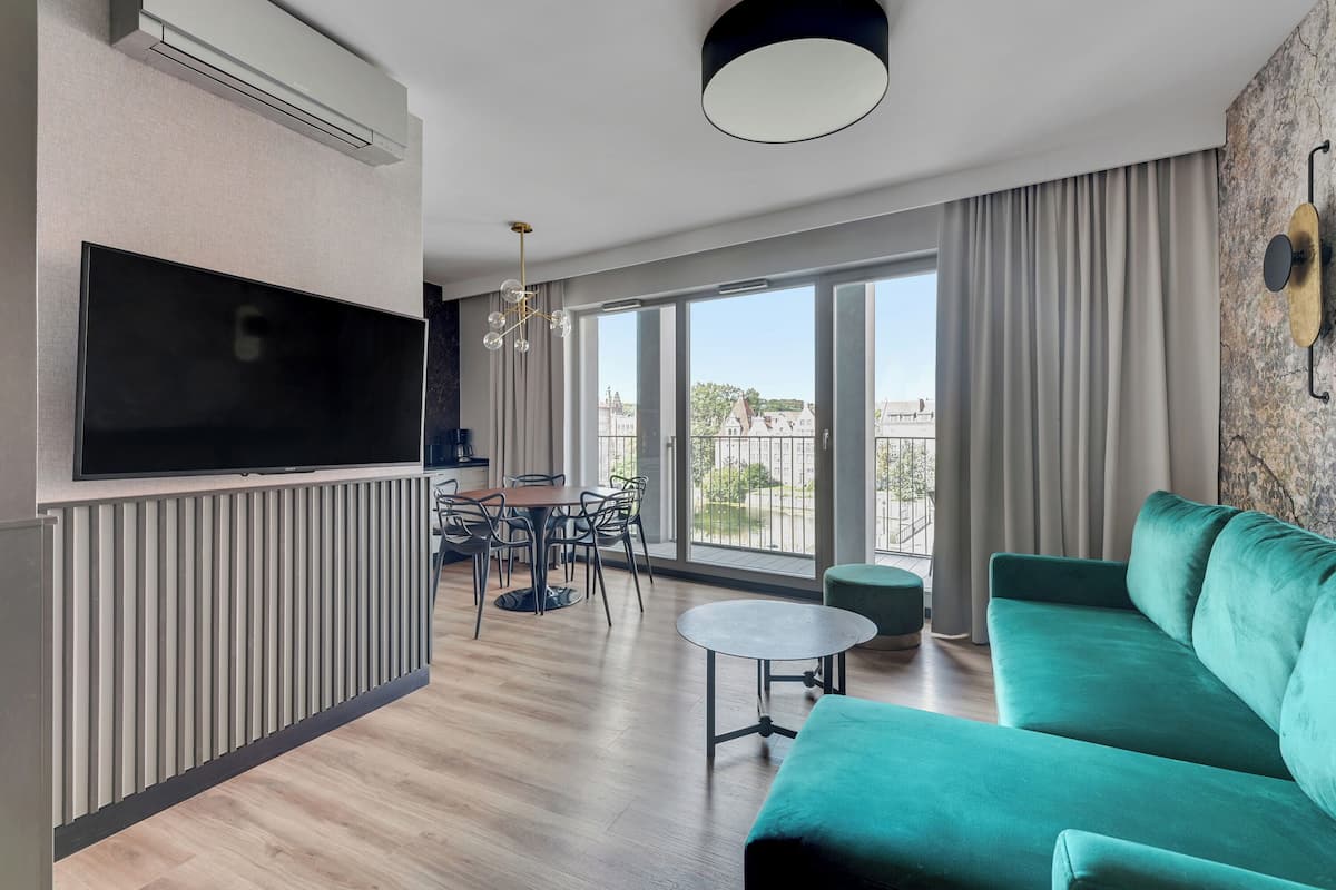 Un salon moderne doté d’un téléviseur à écran plat, d’un canapé vert, d’une table à manger avec des chaises et de grandes fenêtres offrant une vue sur la ville.