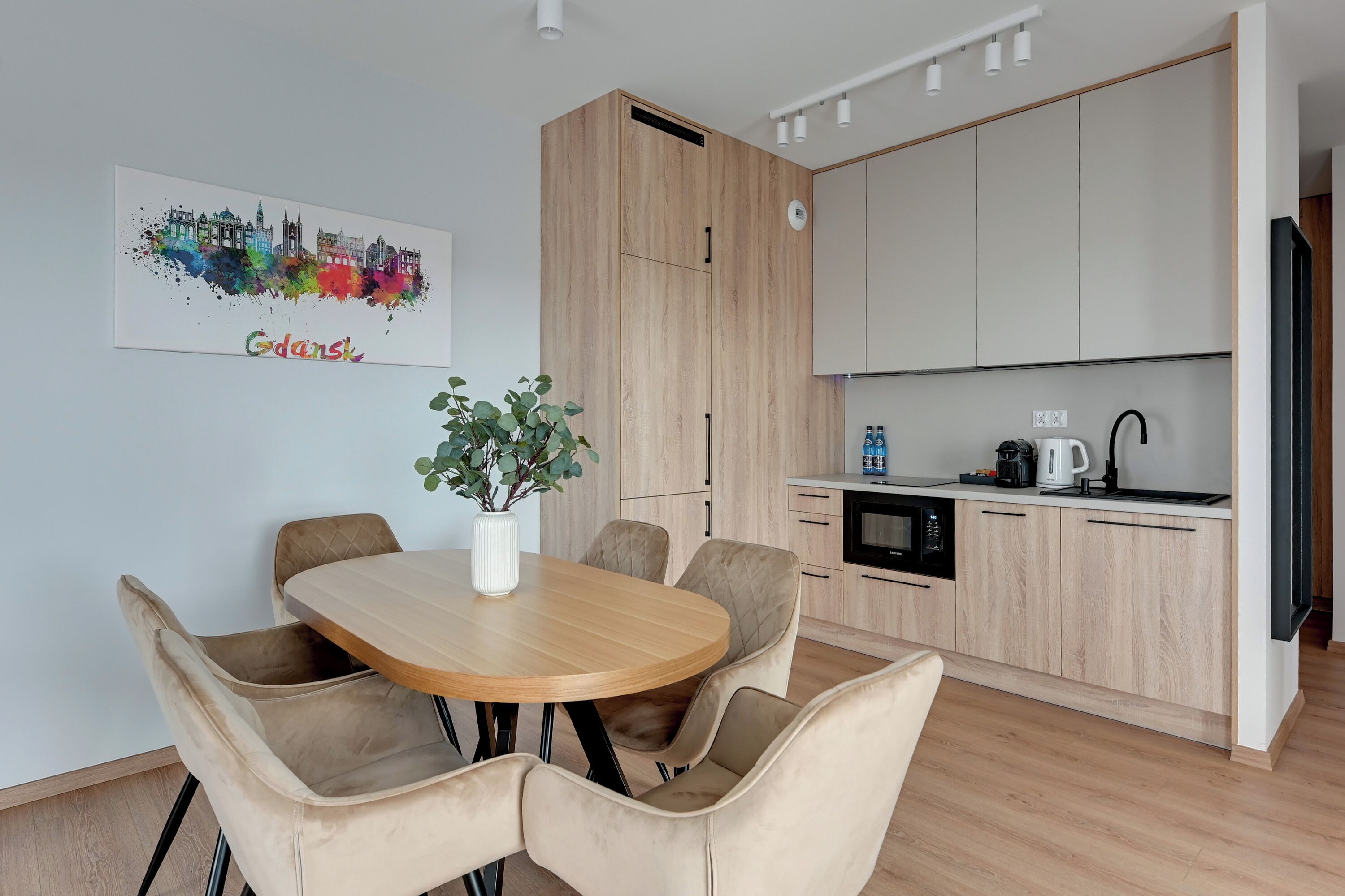 Een moderne keuken met lichte houten kastjes, een eetgedeelte met een houten tafel en gestoffeerde stoelen, en een stadsgezicht kunstwerk aan de muur.