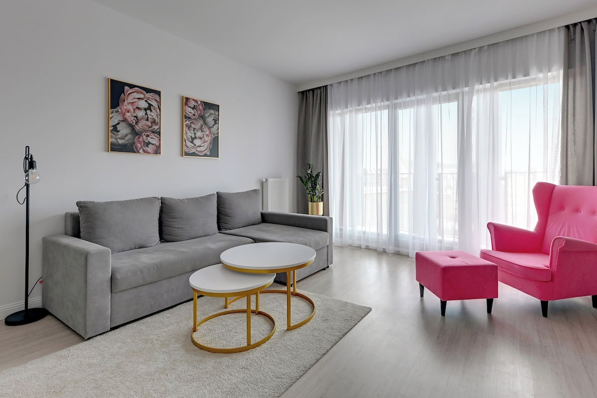 Un salon moderne avec un canapé gris, deux tables rondes, un fauteuil rose et de grandes fenêtres ornées de rideaux légers.
