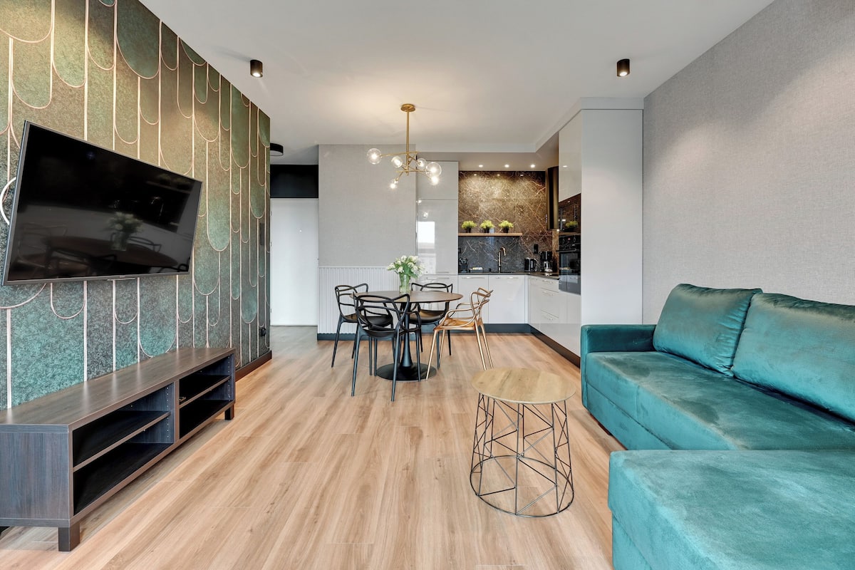 Un salon moderne avec un téléviseur à écran plat, un canapé de couleur turquoise, un coin repas avec une table ronde et une cuisine en arrière-plan.