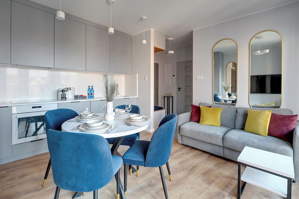 Une cuisine moderne avec des meubles gris, une table à manger entourée de chaises bleues et un canapé gris agrémenté de coussins jaunes et rouges.