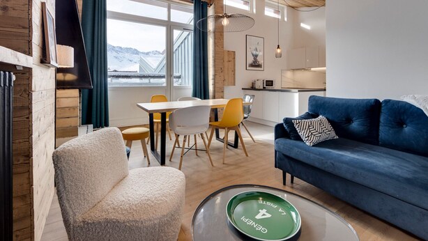 Un intérieur moderne avec un plafond en bois, un coin repas avec une table blanche et des chaises jaunes, un canapé bleu et une vue sur des montagnes enneigées à travers de grandes fenêtres.