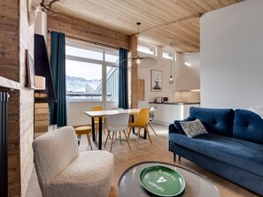 Et moderne interiør med træloft, et spiseområde med et hvidt bord og gule stole, en blå sofa og udsigt til sneklædte bjerge gennem store vinduer.