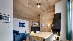 Un salon moderne avec un plafond en bois, une cheminée, une table à manger avec des chaises, un canapé bleu et un téléviseur fixé au mur.