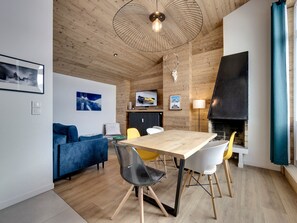 A modern living room with a wooden ceiling, a fireplace, a dining table with chairs, a blue sofa, and a TV mounted on the wall.
