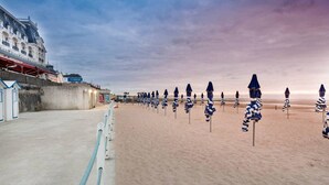A beach with rows of striped beach umbrellas and a building with balconies. - 2-room apartment, 5 people, parking, near the beach (Le Home Varaville)