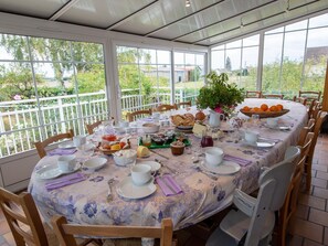 A round table set for breakfast with a floral tablecloth, surrounded by wooden chairs, in a room with large windows offering a view of greenery and buildings. - Room La Loire - Disabled Access, WiFi, Pets Allowed (Saint-Benoît-sur-Loire)