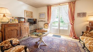 A cozy living room with a wooden ceiling, a television, a fireplace, and a large window with curtains. - Room La Loire - Disabled Access, WiFi, Pets Allowed (Saint-Benoît-sur-Loire)