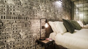 A hotel room with a patterned wallpaper depicting architectural designs, a bedside table with a lamp, and a neatly made bed with pillows. - Elaisa s House - RS (Genoa)