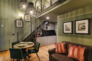 Un coin repas avec une table en bois, des chaises vertes et un canapé marron agrémenté de coussins à rayures. Un escalier mène à une cuisine équipée d’un four à micro-ondes et d’un évier.