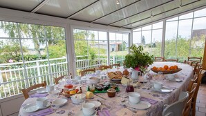 A round table set for breakfast with a floral tablecloth, surrounded by wooden chairs, in a room with large windows offering a view of greenery and buildings. - Charming room with private terrace and wifi in Saint-Benoît-sur-Loire (Saint-Benoît-sur-Loire)