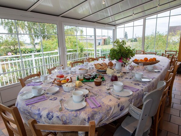 A round table set for breakfast with a floral tablecloth, surrounded by wooden chairs, in a room with large windows offering a view of greenery and buildings. - Charming room with private terrace and wifi in Saint-Benoît-sur-Loire (Saint-Benoît-sur-Loire)