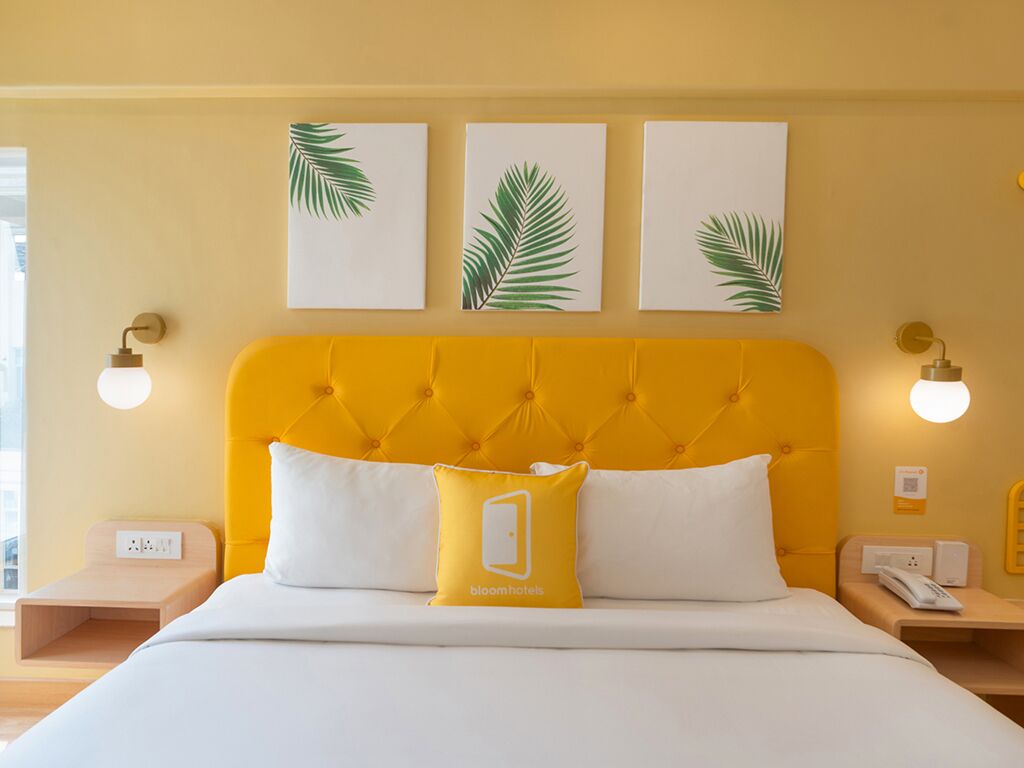 Una habitación de hotel con cabecera amarilla, ropa de cama blanca y tres piezas de arte con hojas de palma.