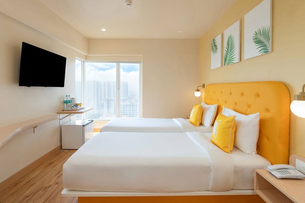 A hotel room with two beds, a flat-screen TV, a desk with a lamp, and a window with a city view.