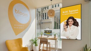 A modern interior with a yellow chair, a wooden table, and a large screen displaying a person. - Bloom Hotel Saki Vihar (Mumbai)