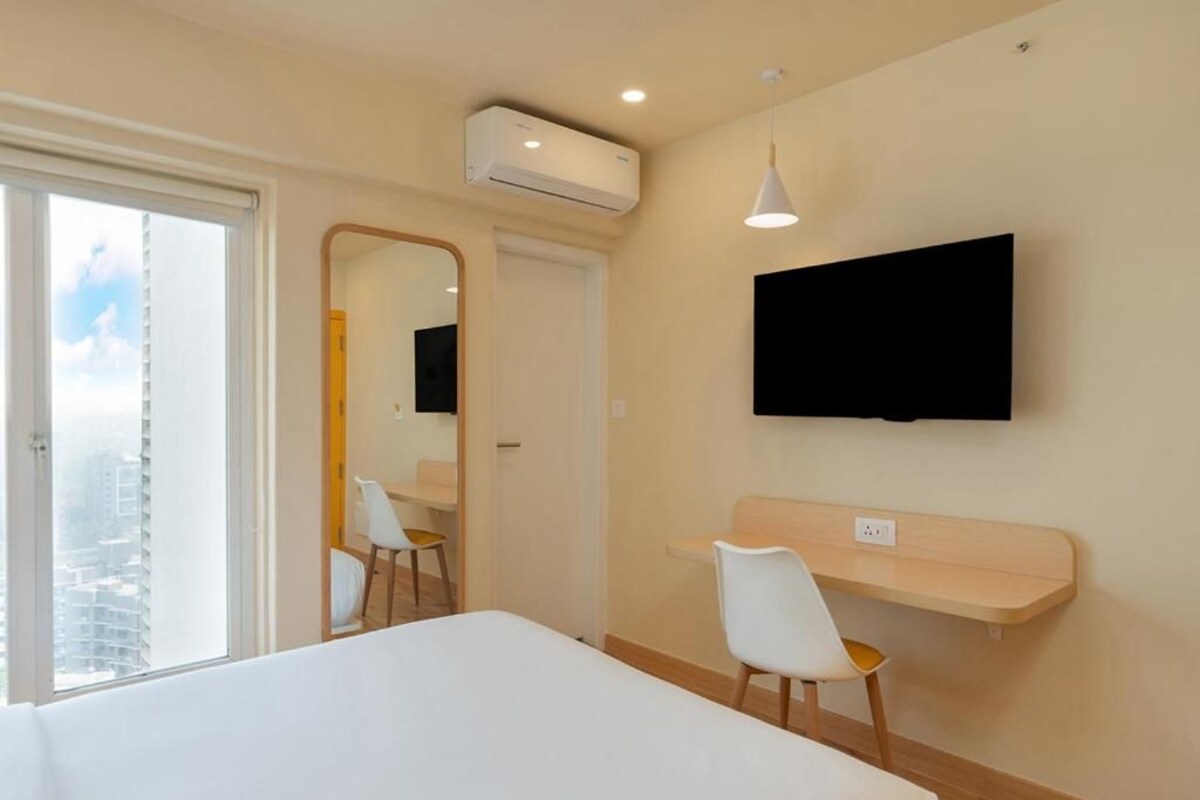 A modern hotel room with a bed, a TV mounted on the wall, a desk with a chair, and a window with a view of buildings.