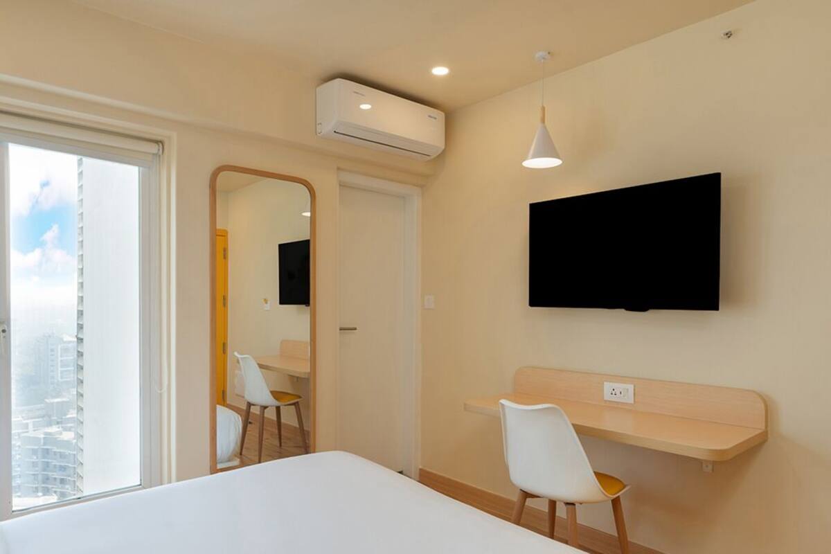A hotel room with a bed, a desk, a chair, a TV mounted on the wall, and an air conditioning unit.