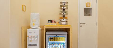 A compact room with a water dispenser, a mini-fridge stocked with beverages, a small snack bar, and a door with a 'Start Only' sign.