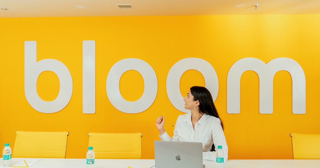 Un salón de conferencias con un cartel grande que dice 'bloom' en la pared, una mesa blanca y sillas amarillas.